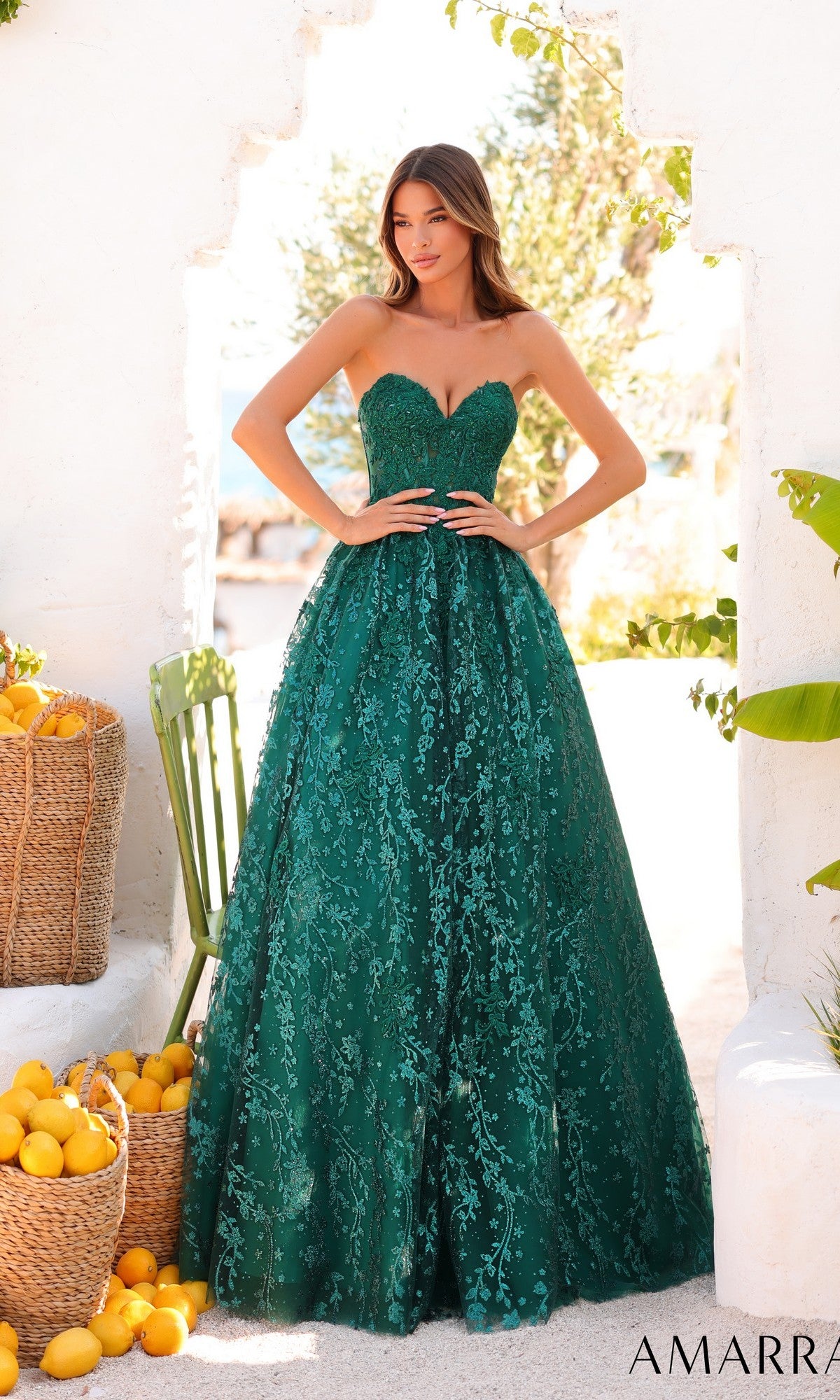 A woman in the Amarra 89427 long prom dress, featuring a strapless lace corset bodice, stands outside near baskets of lemons and white stone walls.